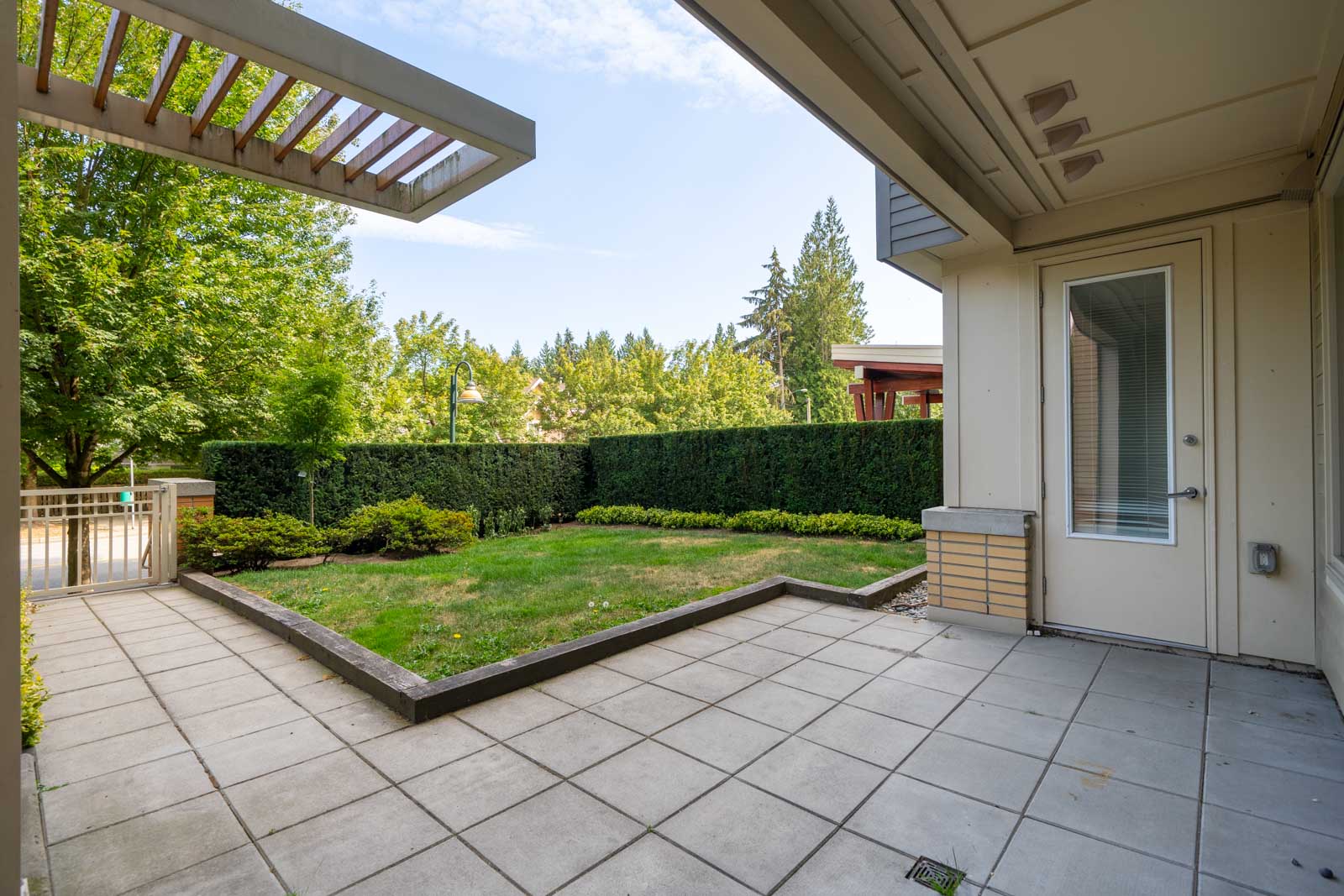 Covered patio with tiled floor overlooks a small grassy yard bordered by shrubs, trees, hedge, and wooden fence in North Vancouver.