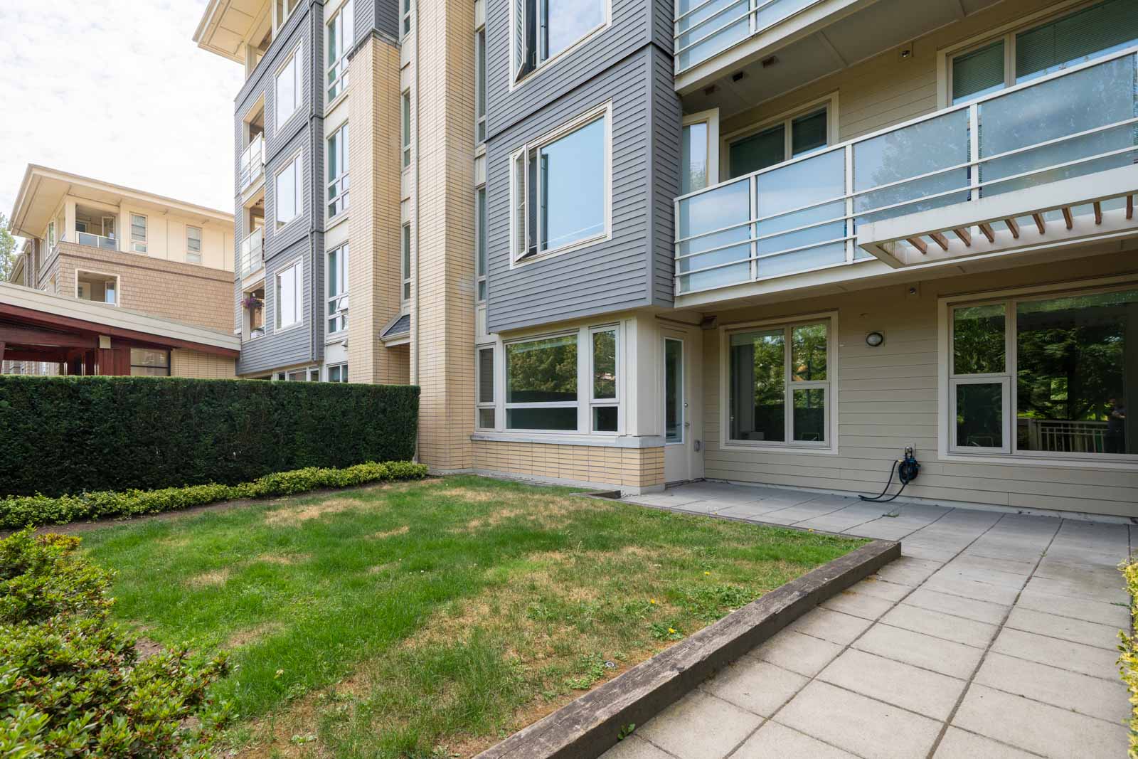 Ground-floor apartment with patio and small lawn, bordered by hedges, in a modern building on Mountain Hwy, North Vancouver.