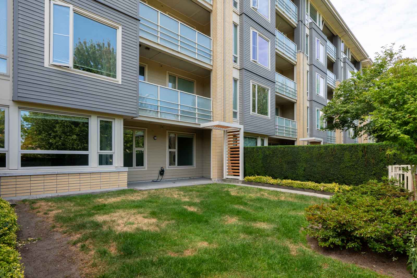 Ground-level patio and grassy yard at a modern apartment with large windows, balconies, and hedges in North Vancouver.