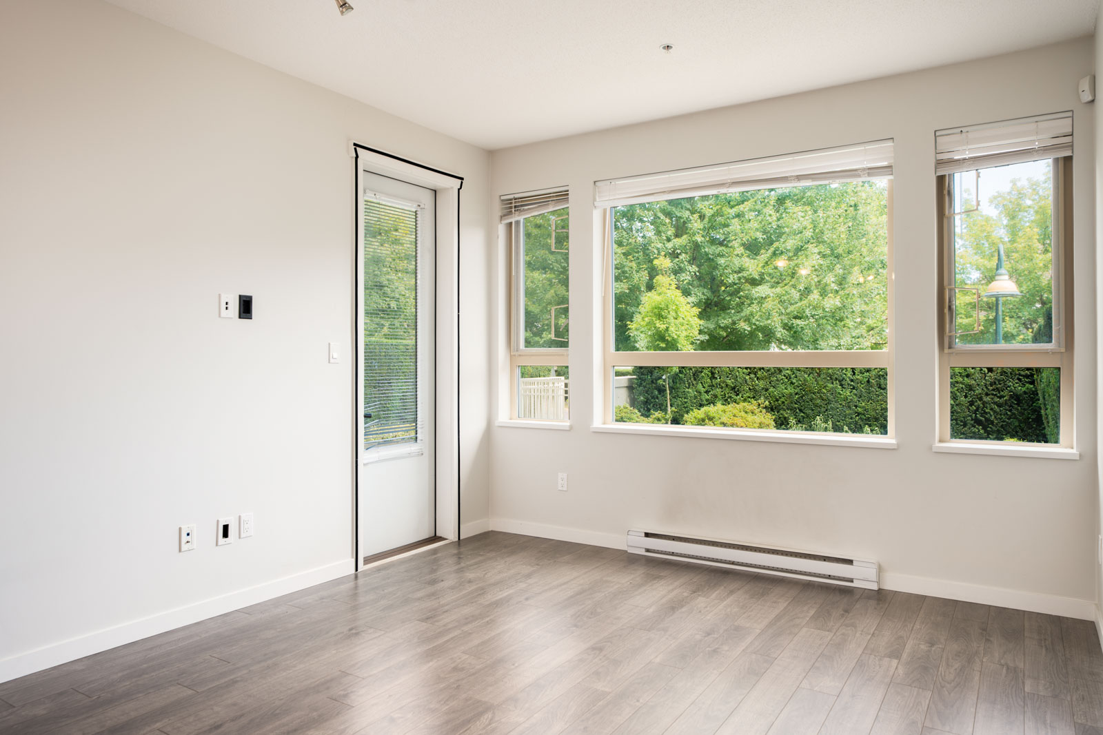 Empty room with light gray walls, large windows overlooking green trees, gray wood floors, baseboard heater, door with blinds in North Vancouver near Mountain Hwy.