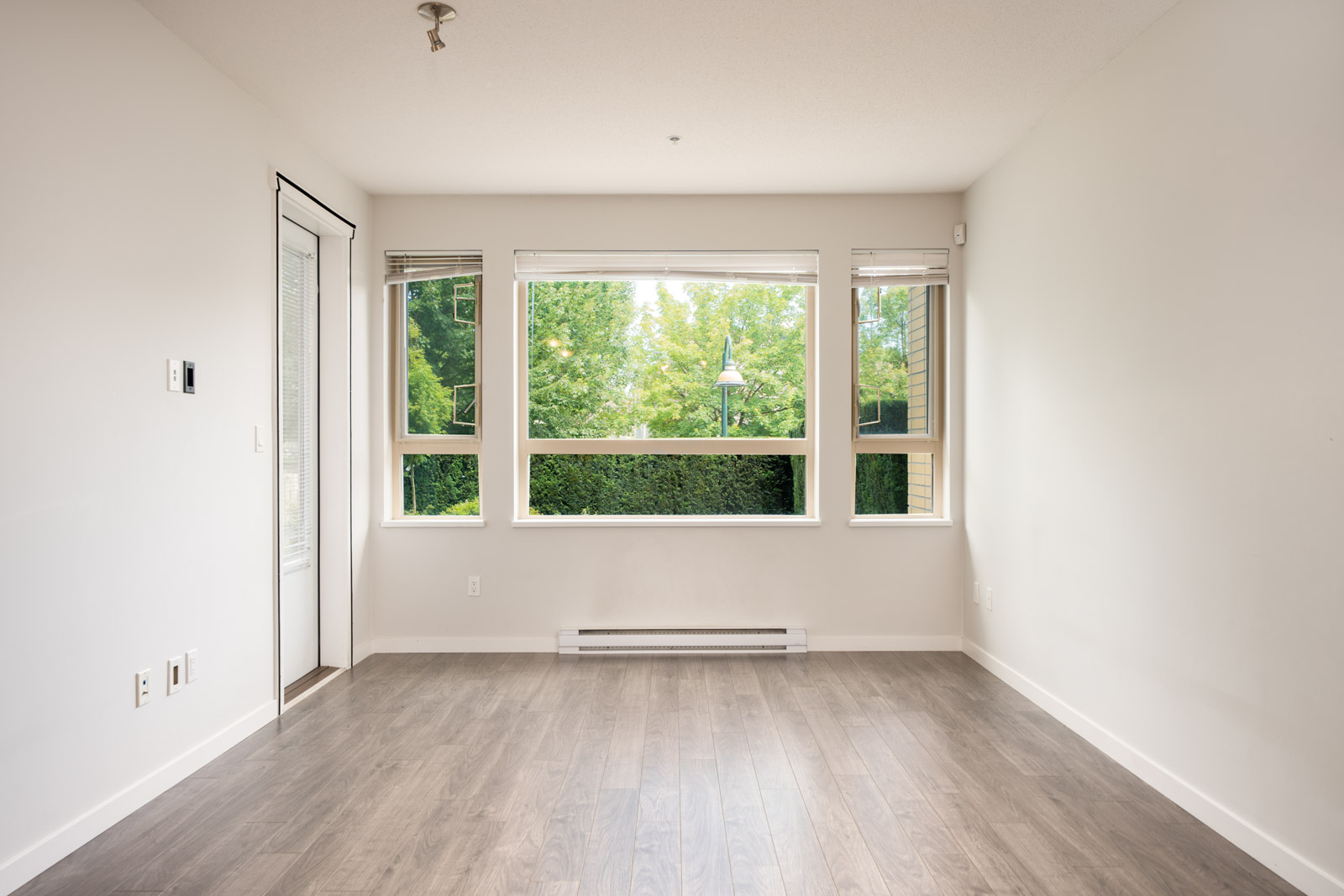Bright, empty room in North Vancouver near Mountain Hwy with pale walls, large windows facing trees, wood floors, and a glass door.