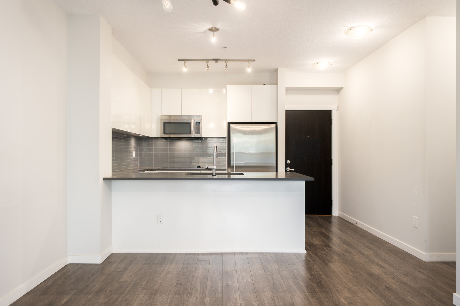 Modern North Vancouver kitchen with white cabinets, stainless steel appliances, gray backsplash, and wood floors, seen from living area.