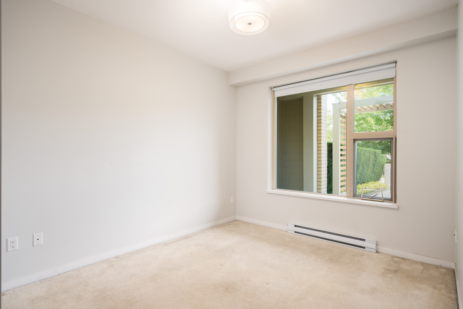 Empty room with beige carpet, white walls, a window with partially open blinds, and ceiling light in North Vancouver near Mountain Hwy.