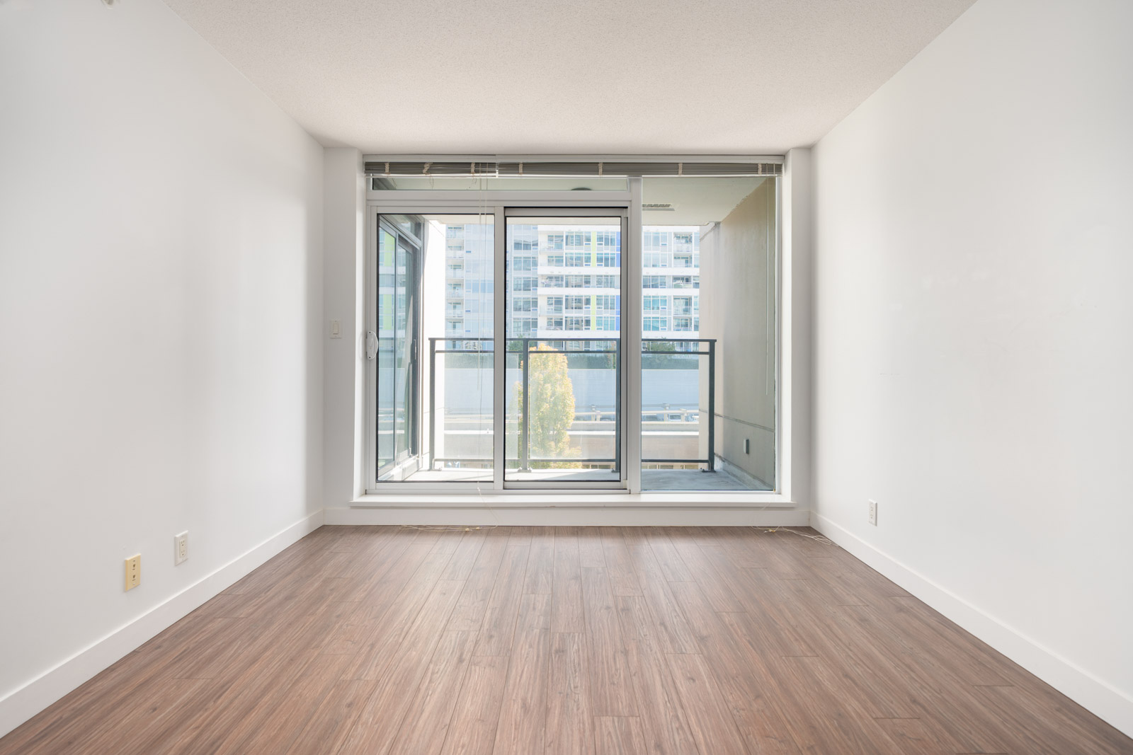 Empty room with white walls, wood floors, and large windows facing a balcony and apartments on Saba Road in Richmond.