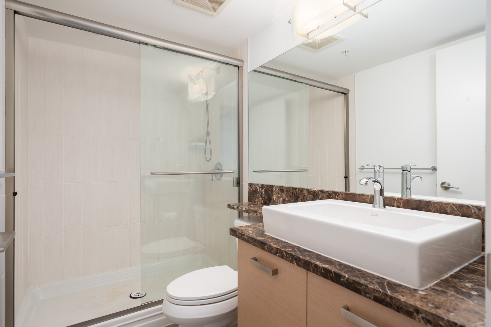 Modern bathroom on Saba Road in Richmond with glass shower, large mirror, white sink, marble counter, and light wood cabinets.
