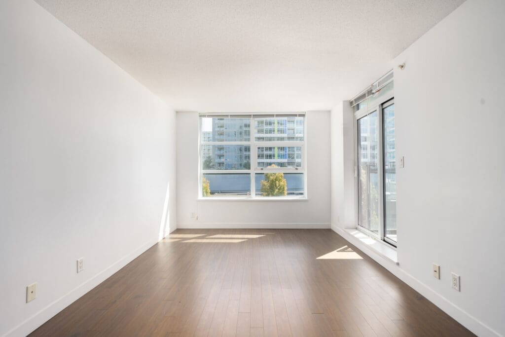 Bright, empty room with white walls, wooden floor, large window and glass door. Sunlight and city view; 6th Floor, Richmond.