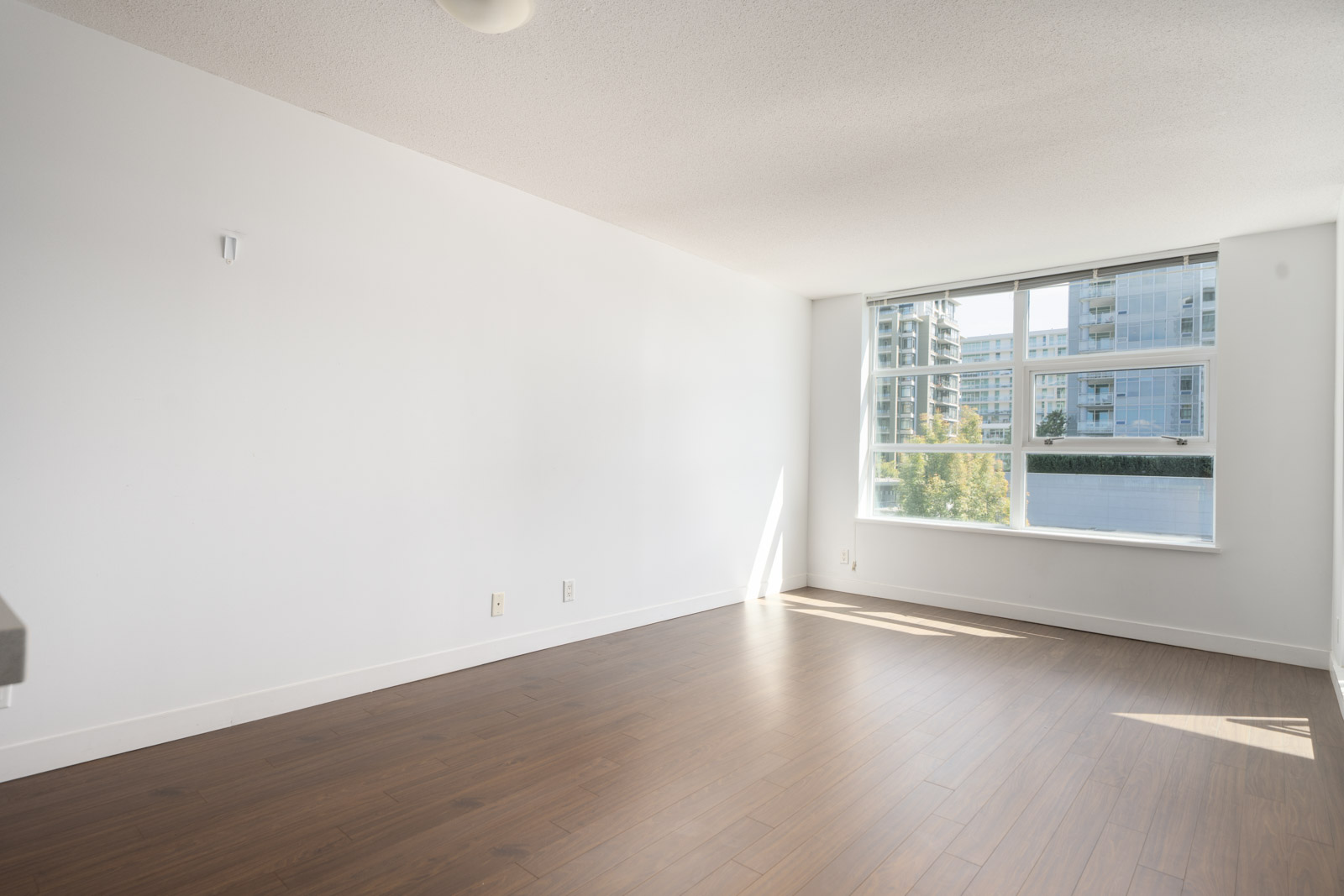 Bright, empty room with white walls, brown wood floor, and large window overlooking Saba Road buildings and trees in Richmond.