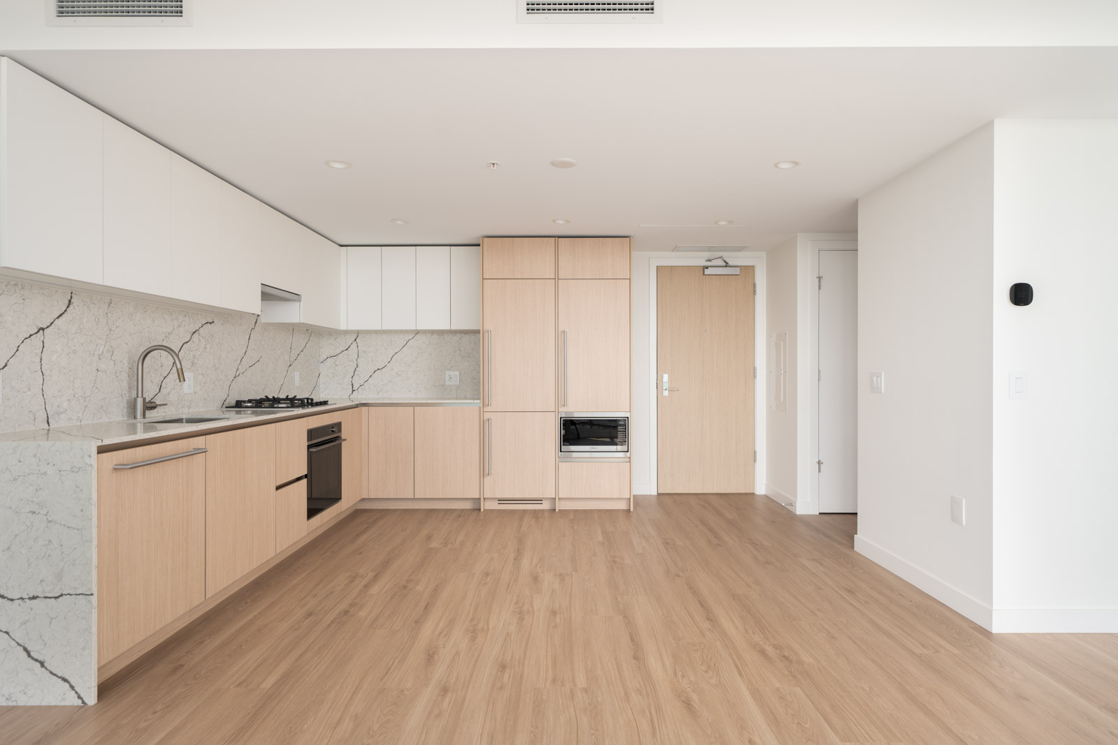 Modern, unfurnished kitchen with light wood cabinets, built-in appliances, marble backsplash, and light wood flooring in a bright, open space.