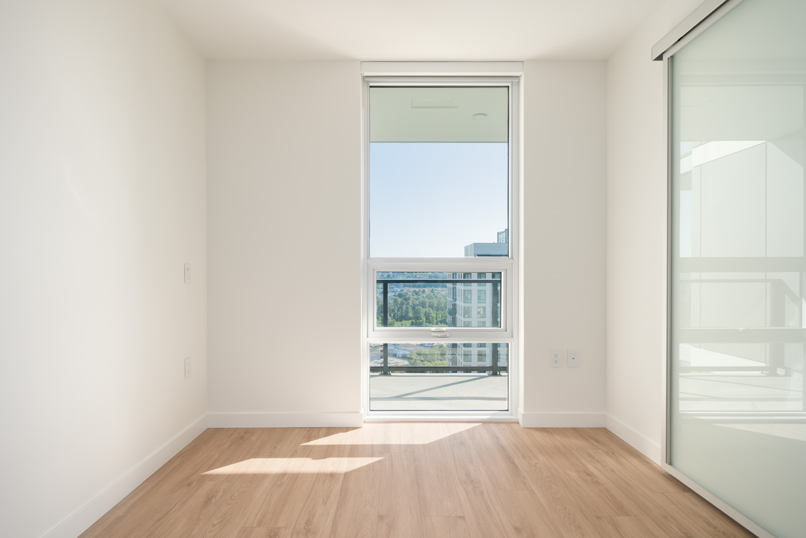 Bright empty room with light wood floor, white walls, floor-to-ceiling window, and glass sliding door; view of cityscape and greenery outside.