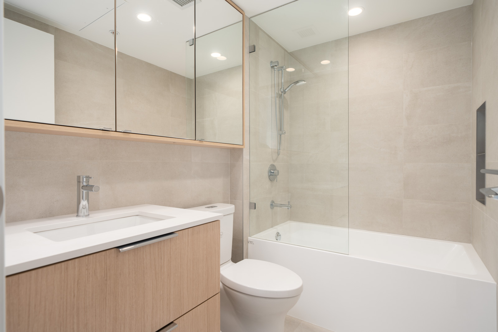 Modern bathroom with light wood vanity, white sink, toilet, and a bathtub with glass shower enclosure, featuring beige tiled walls and a mirrored cabinet.