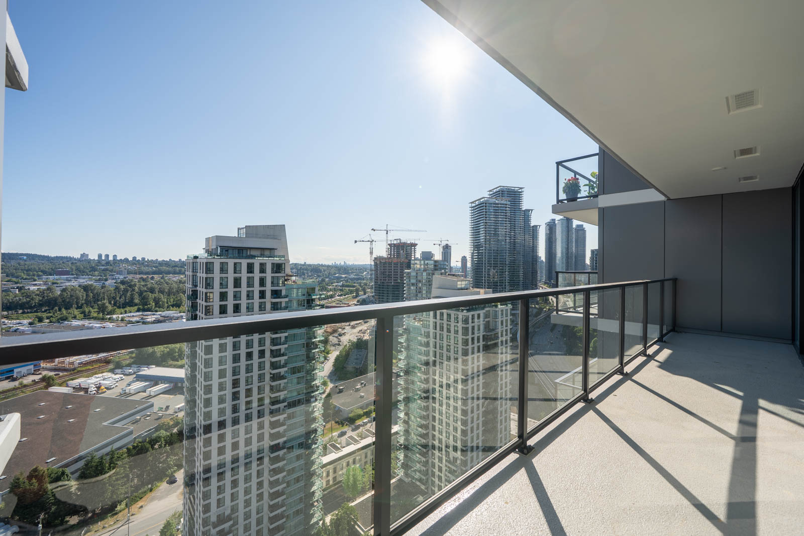 High-rise balcony with glass railing overlooking city buildings, construction cranes, and a clear blue sky with the sun shining.