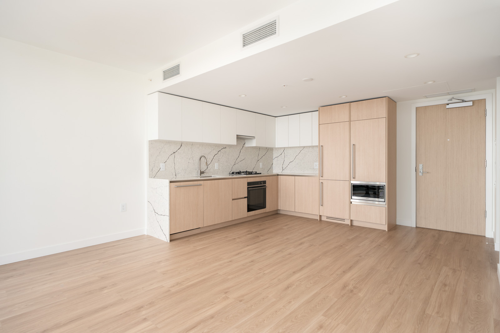 Modern, unfurnished kitchen with light wood cabinets, built-in appliances, marble backsplash, and light wood flooring in a bright, empty room.