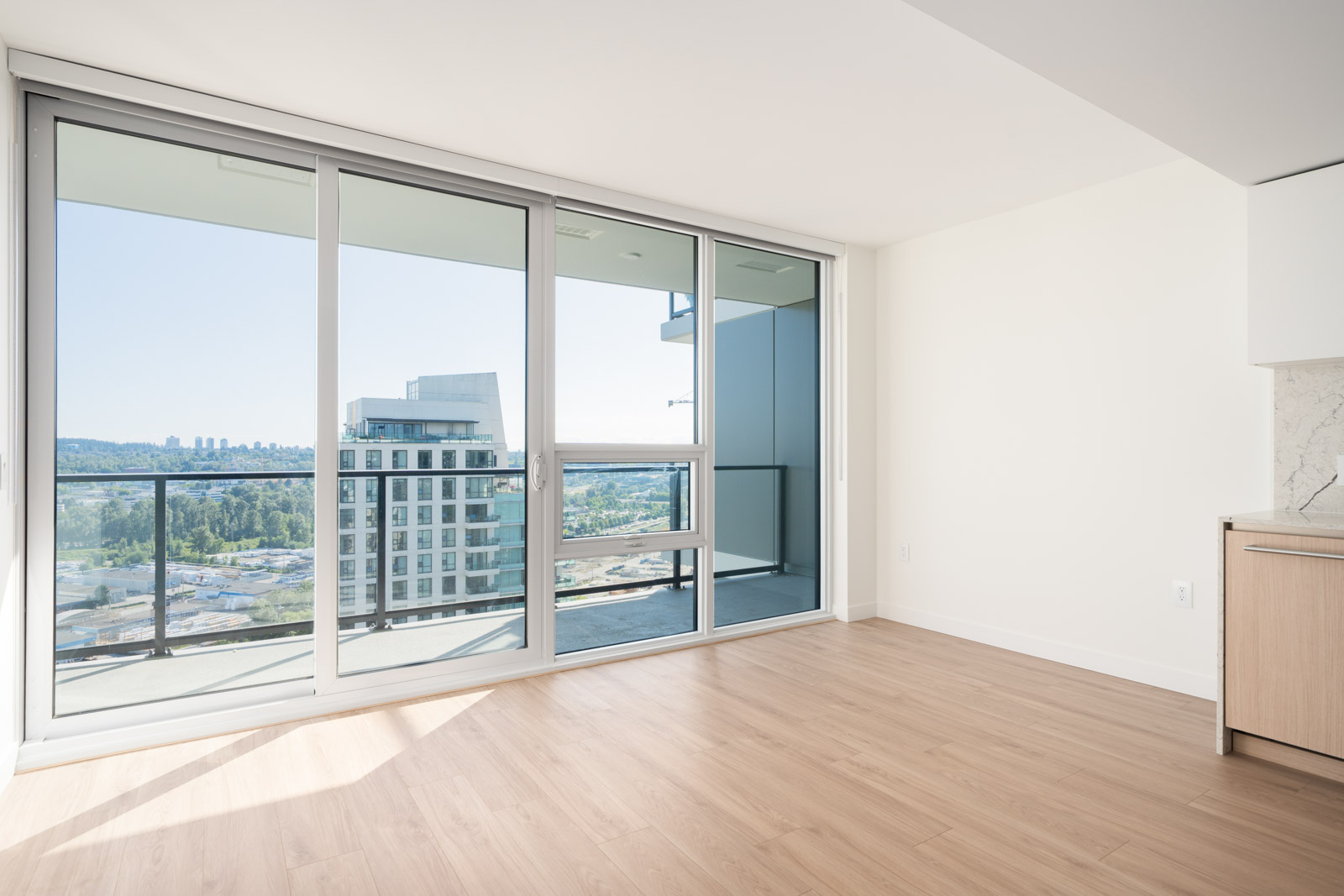 Bright, empty room with light wood flooring, floor-to-ceiling windows, and a balcony overlooking city buildings and greenery on a sunny day.
