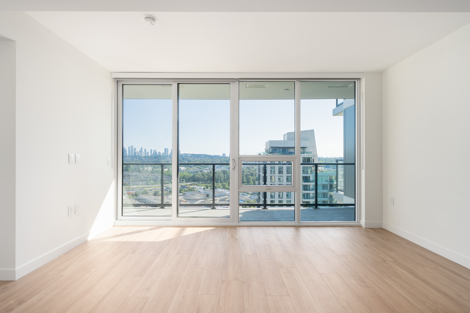 Empty modern apartment room with light wood floor, white walls, and large floor-to-ceiling windows opening to a balcony with city and skyline views.