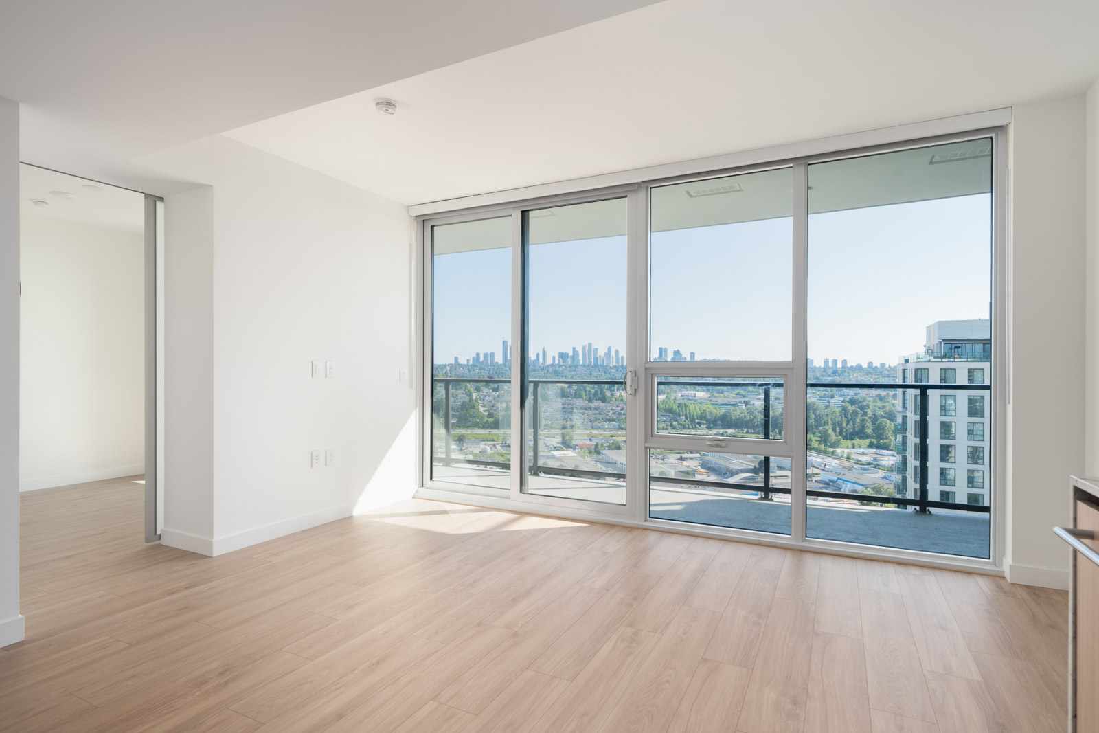 Bright, empty apartment room with light wood flooring, large floor-to-ceiling windows, a balcony, and a city skyline view in the distance.