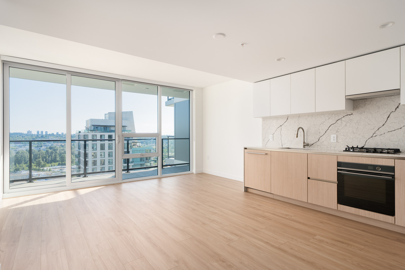 Modern, unfurnished apartment with light wood flooring, minimalist kitchen, marble backsplash, and large floor-to-ceiling windows leading to a balcony with a city and park view.