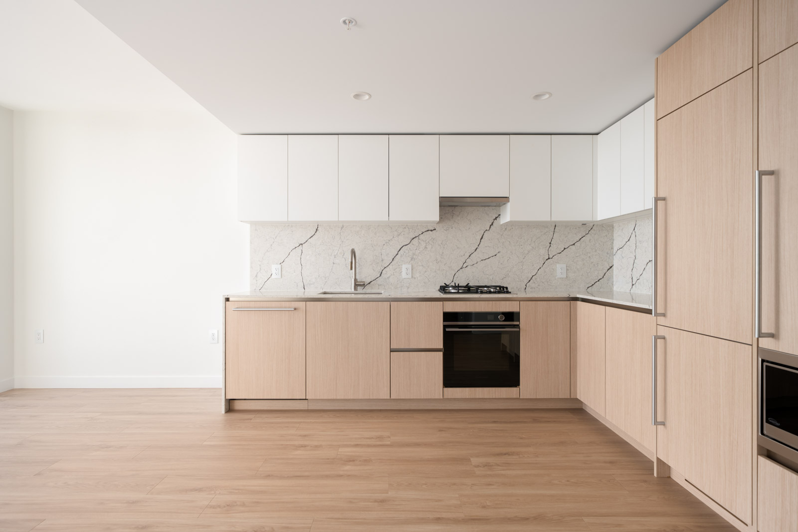 Modern kitchen with light wood cabinets, white upper cabinets, marble-patterned backsplash, built-in oven, stovetop, sink, and hardwood flooring.