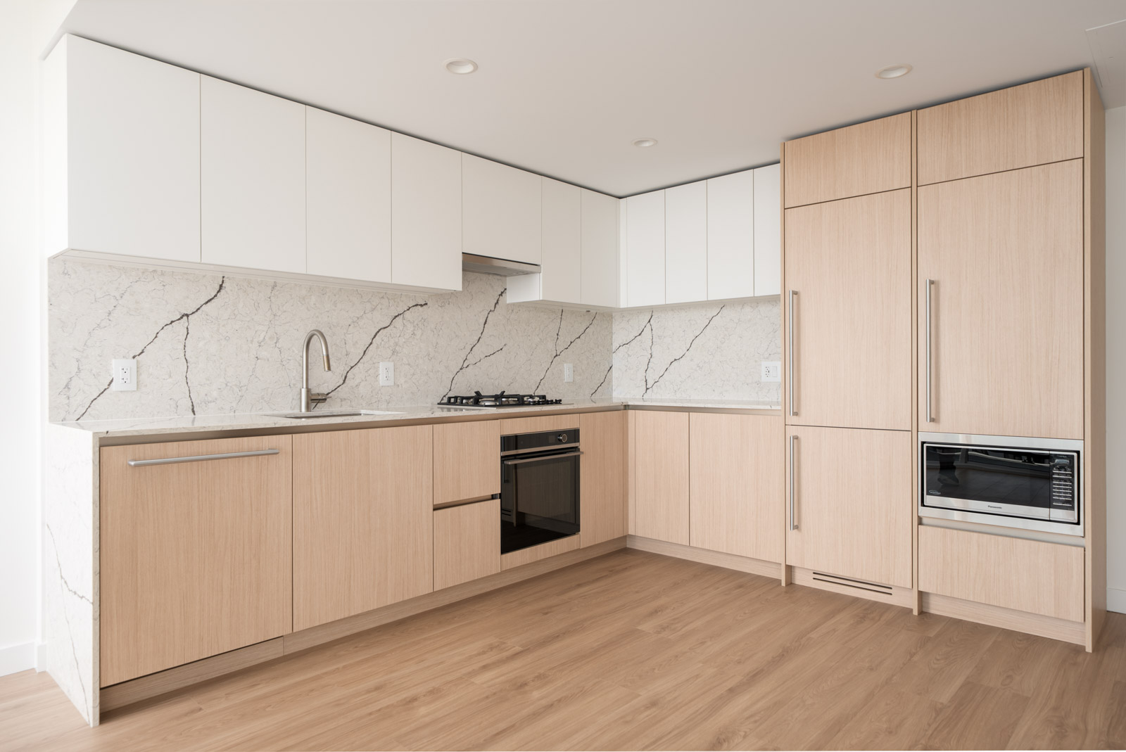 Modern kitchen with light wood cabinets, white upper cabinets, marble backsplash, built-in oven, stovetop, refrigerator, and microwave, all set on a light wood floor.