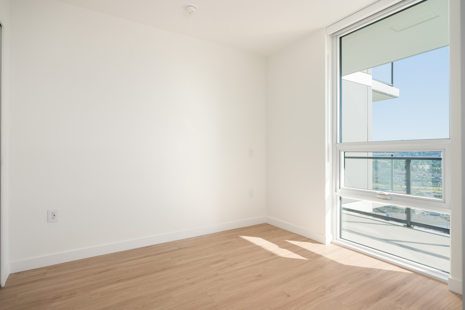 Empty, unfurnished room with white walls, light wood flooring, and a large window letting in natural light and providing a view of a balcony and cityscape.