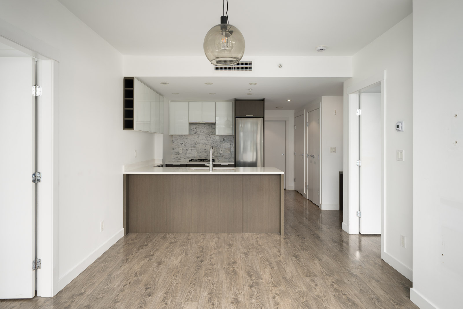 Modern kitchen with a large island, white cabinets, stainless steel appliances, marble backsplash, and wood flooring in a bright, empty apartment.