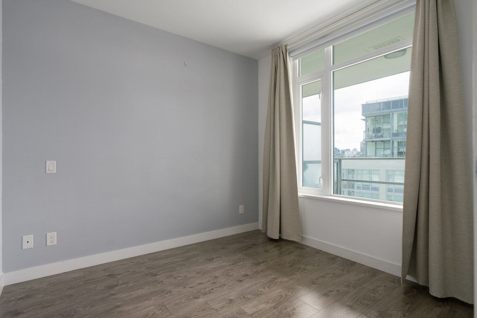 Unfurnished room with light gray walls, large window with beige curtains, and hardwood flooring. A modern building is visible outside the window.
