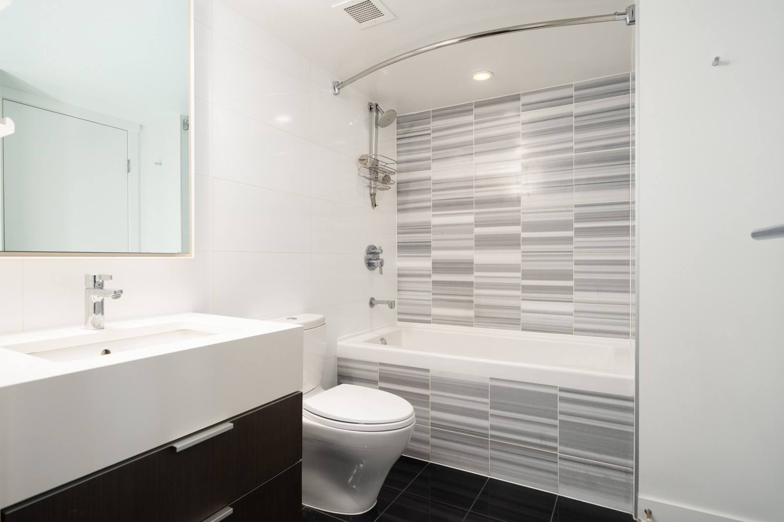 Modern bathroom with a sink, toilet, and bathtub. The bathtub has a gray and white striped tile wall, and the floor is covered with black tiles.