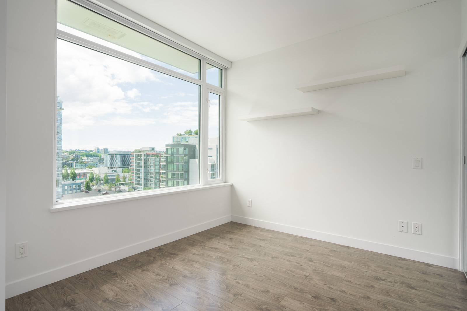 Empty, unfurnished room with white walls, two floating shelves, large window overlooking urban buildings, and wood-style flooring.