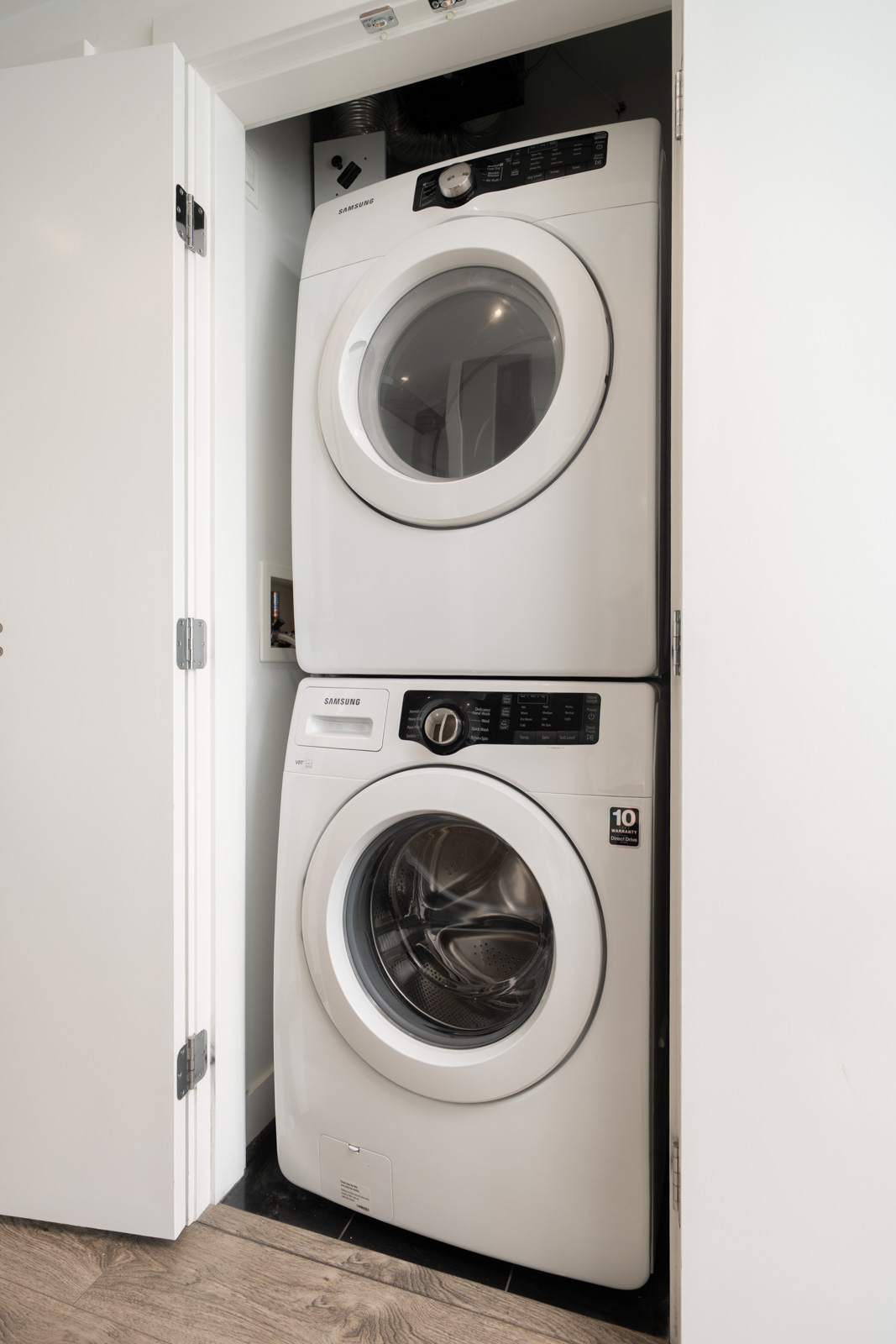 Stacked white Samsung washer and dryer units inside a small closet with open white doors, set on a wood floor.