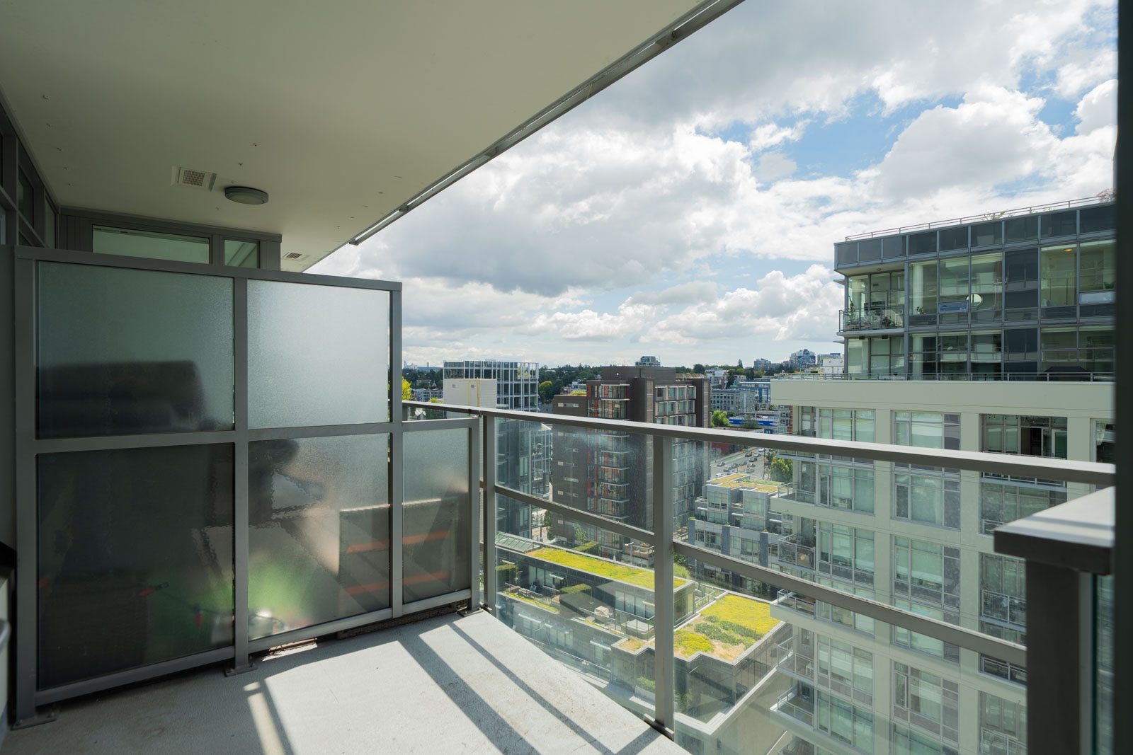 A modern apartment balcony with glass railing and privacy screen overlooks nearby residential buildings under a partly cloudy sky.