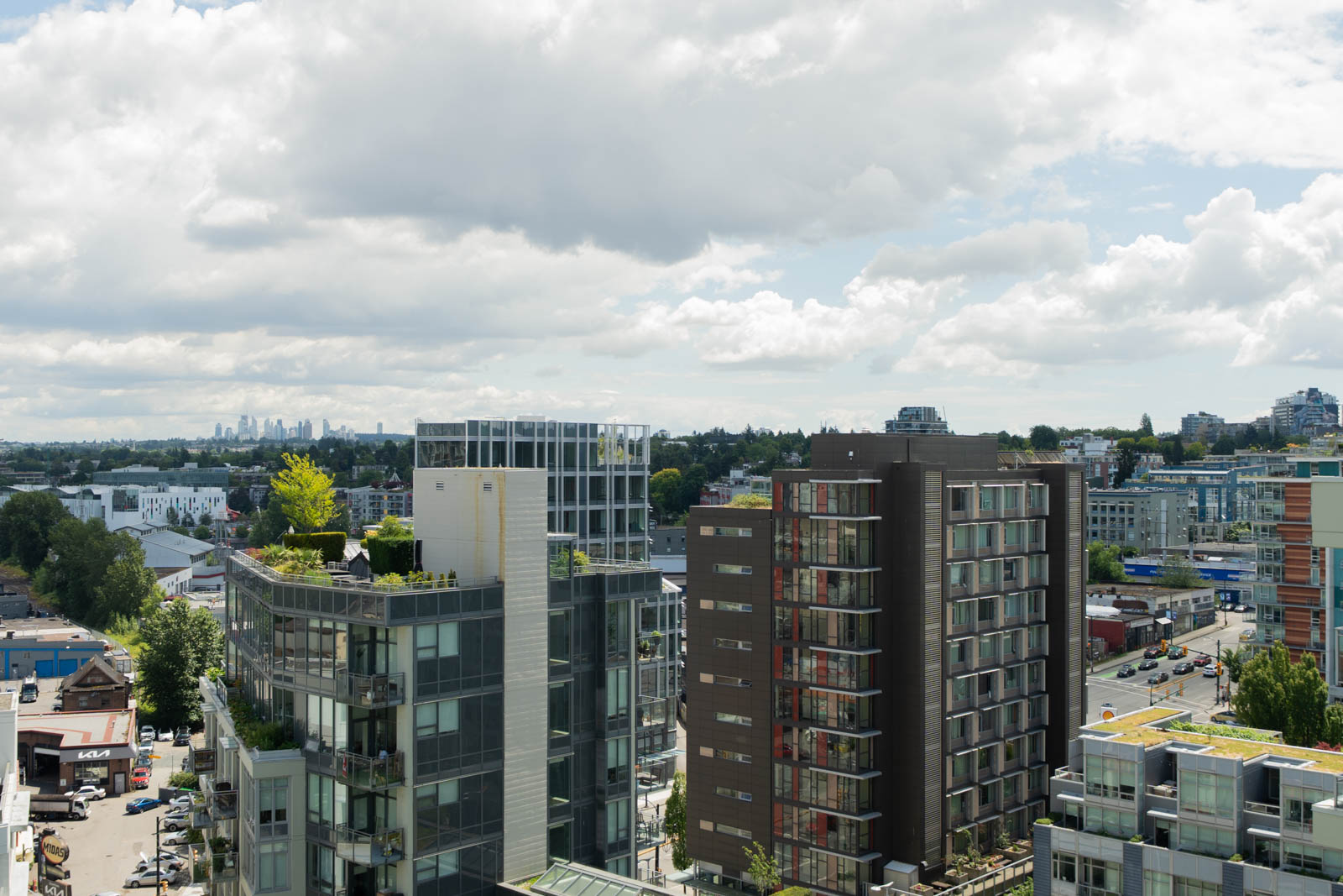 Several mid-rise apartment buildings with rooftop gardens under a partly cloudy sky, with a mix of residential and commercial areas in the background.