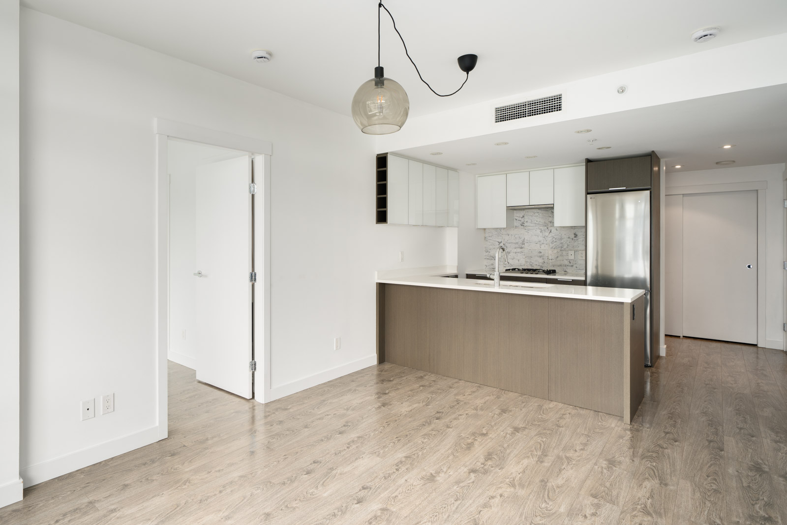 Modern kitchen with light wood flooring, white cabinets, stainless steel appliances, and a hanging light fixture. An open doorway leads to another room.
