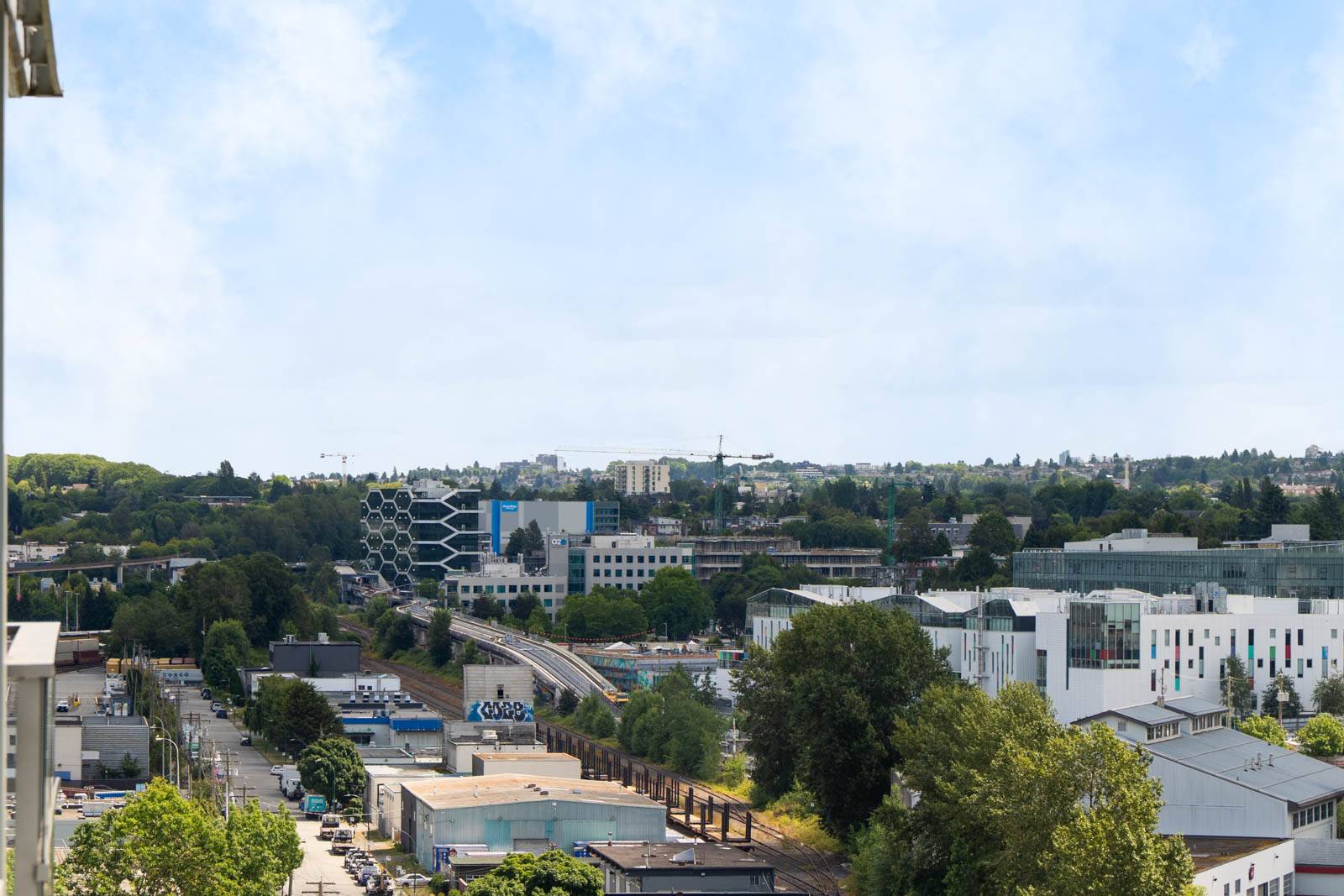 Urban landscape with modern buildings, railway tracks, trees, and a partly cloudy sky in the background.