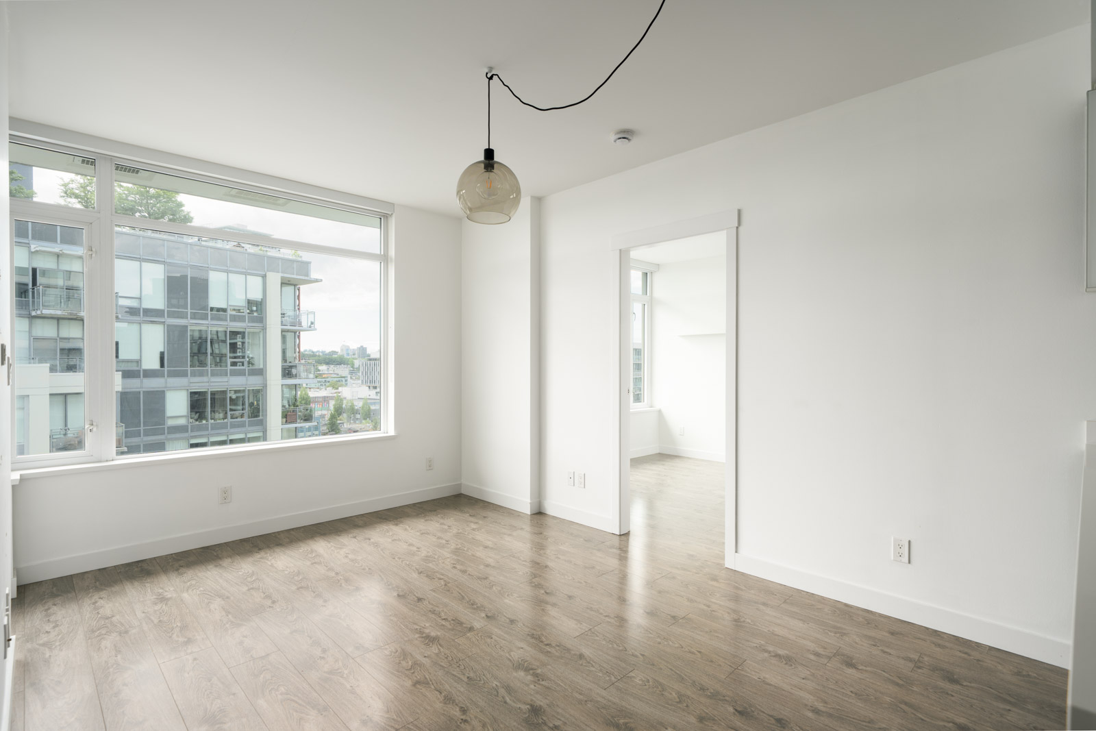 Unfurnished room with large windows, white walls, wood flooring, and a hanging light fixture; doorway leads to another bright room.