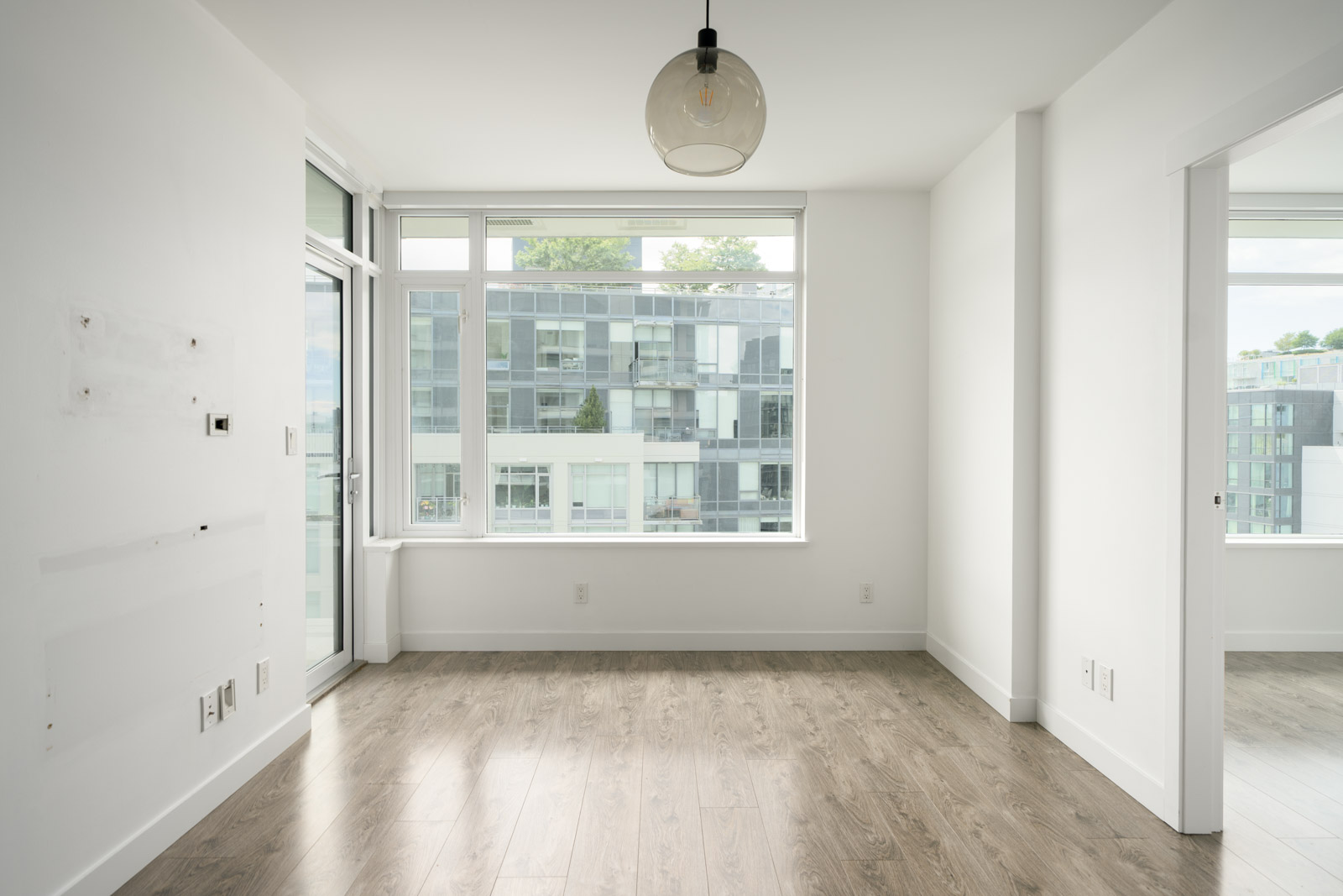 Empty modern apartment room with light wood flooring, white walls, large window, and glass door overlooking a contemporary building.