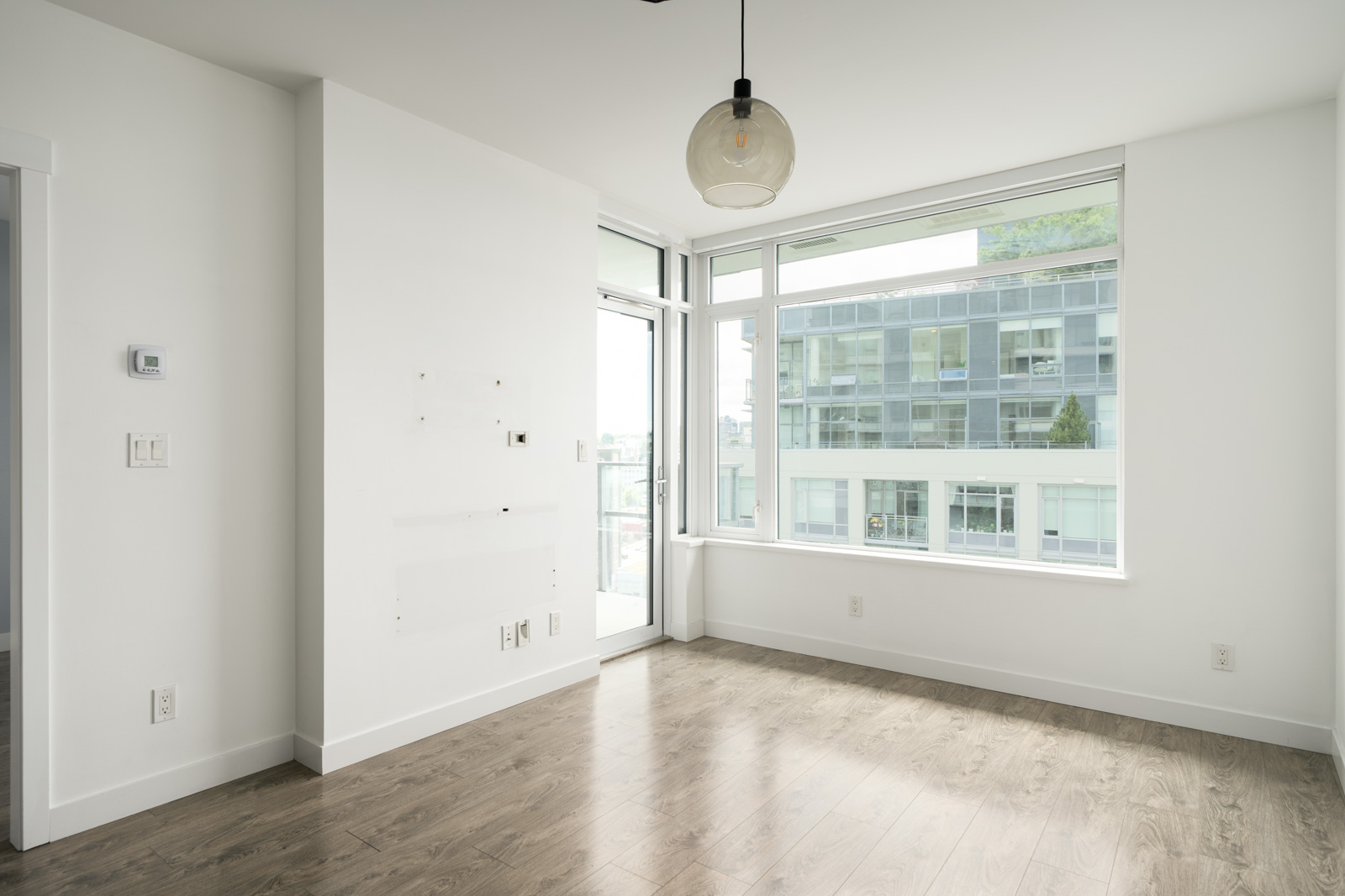 Empty, modern apartment room with white walls, large windows, wood flooring, a ceiling light fixture, and a small balcony visible outside.
