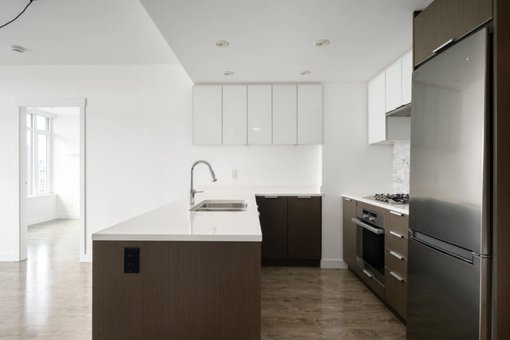 Modern kitchen with brown and white cabinets, built-in oven, gas cooktop, stainless steel refrigerator, white countertops, and wood flooring. Bright natural light from adjacent room.