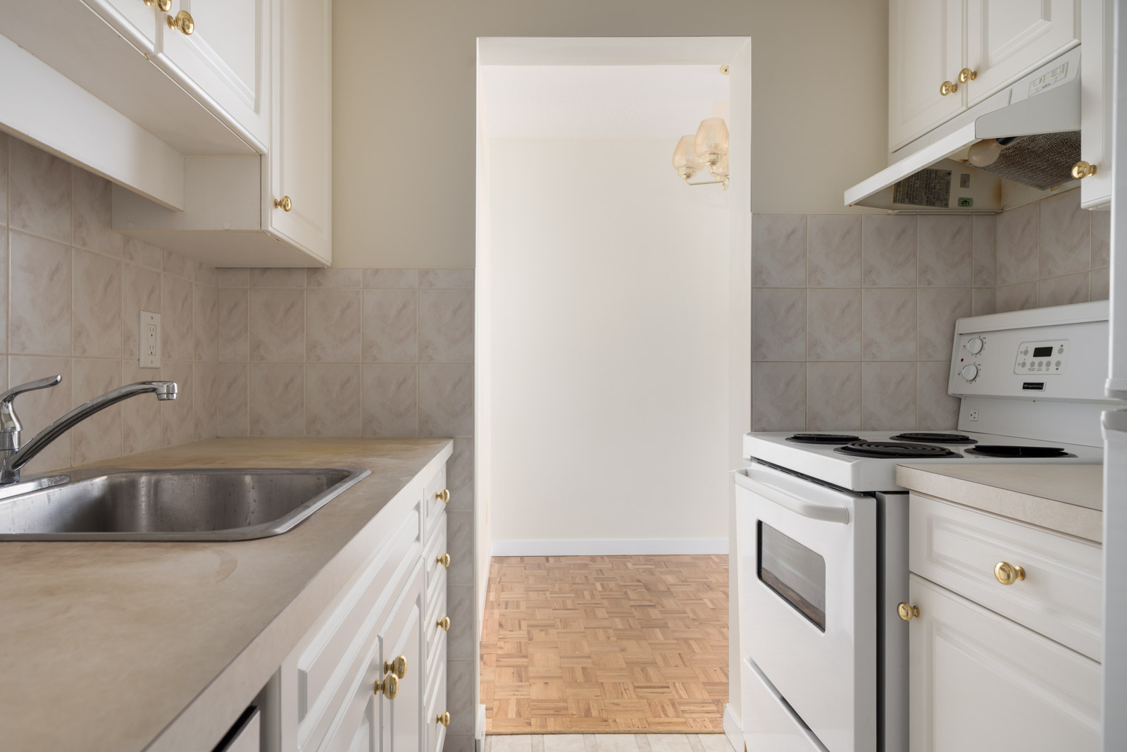 Narrow white kitchen in Vancouver with electric stove, sink, gold hardware, opening to a dining area with parquet floor and ceiling light.