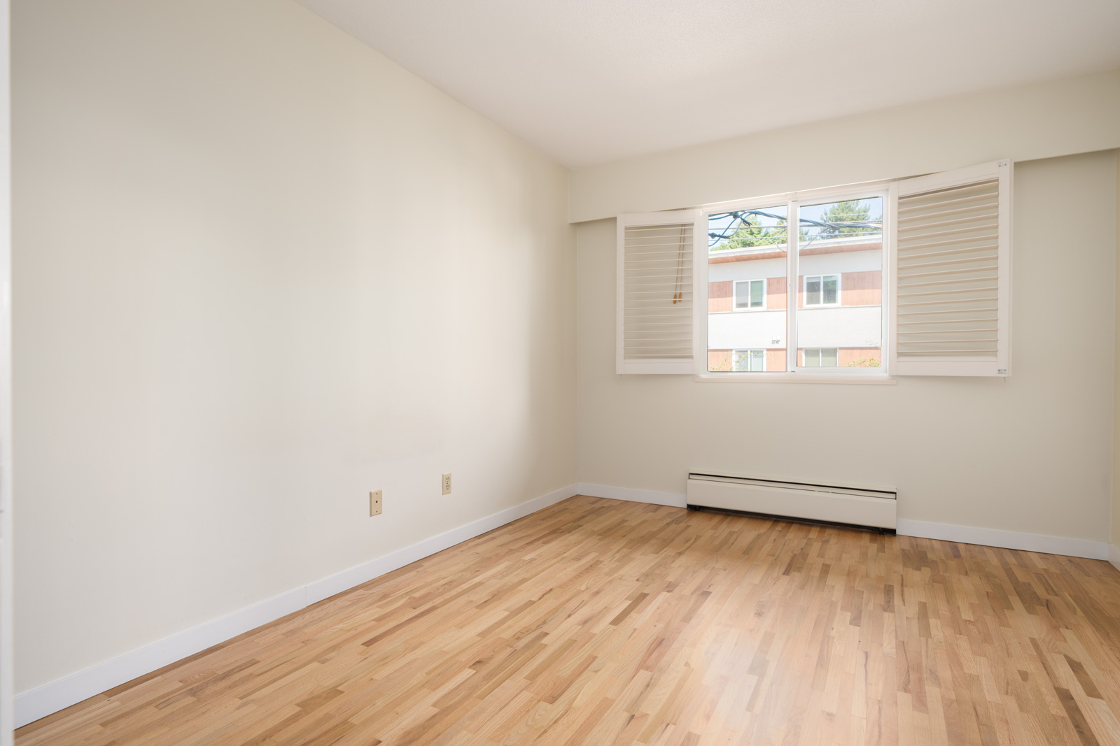 Empty room with light wood floors, beige walls, large shuttered window, and baseboard heater beneath it on W 2nd Ave in Vancouver.