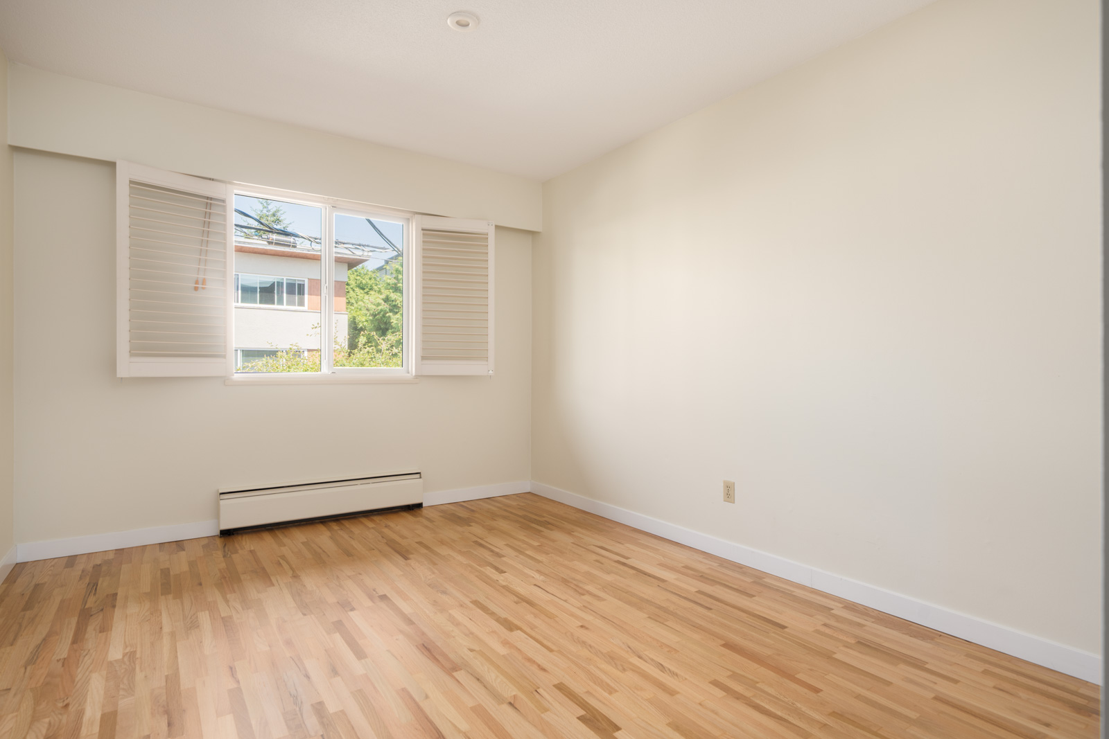 Empty room with pale walls, wood floors, a window with white shutters, and a baseboard heater on W 2nd Ave, Vancouver.