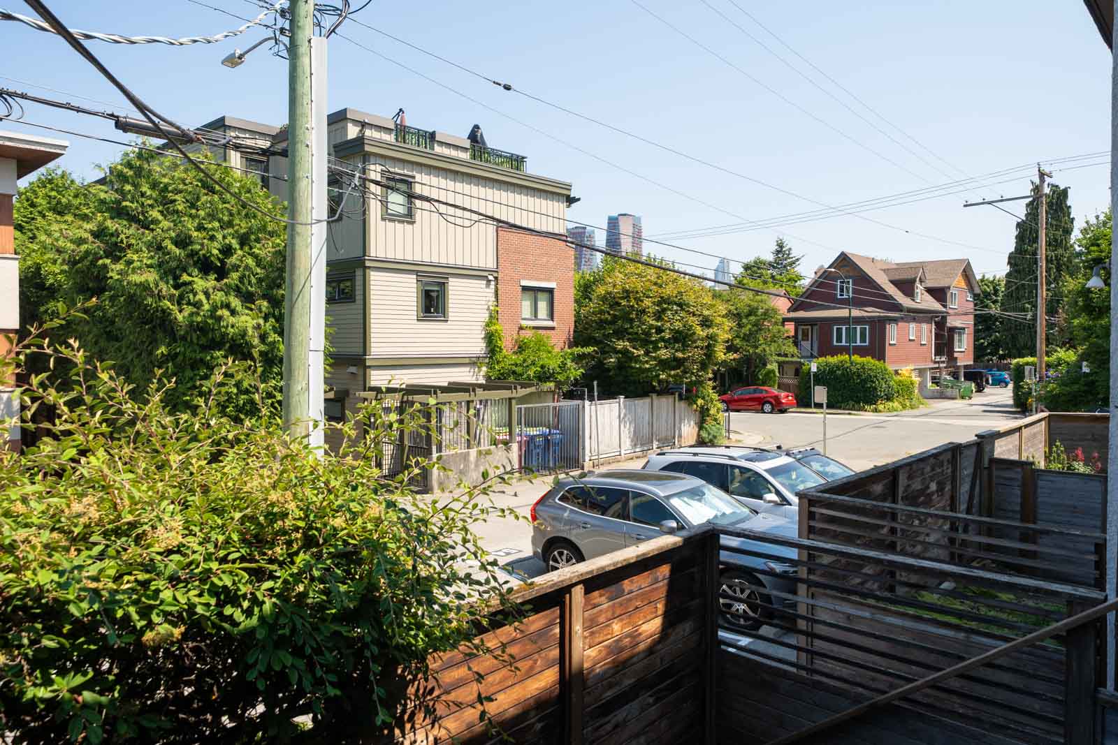 Parked cars along a residential street, wooden fence in front, houses, trees, and power lines under clear sky in Vancouver.