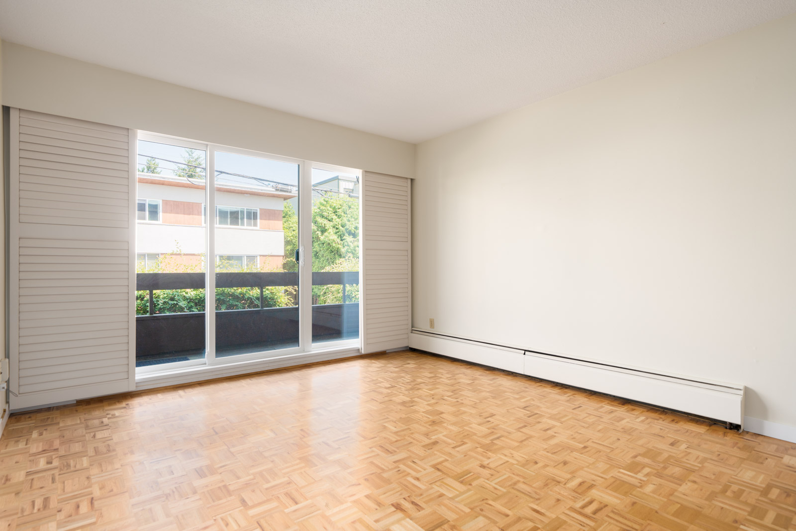 Empty room with light parquet floors, large glass doors, white walls, and balcony view of greenery and apartments in Vancouver.