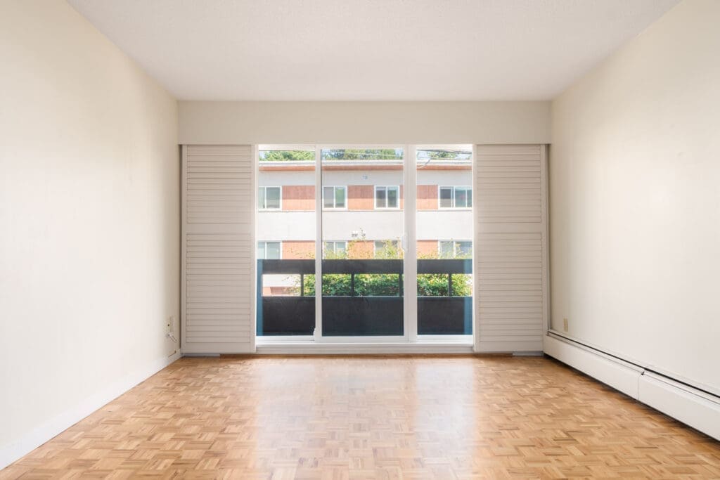 Empty room with light walls, parquet floors, and a large window with sliding shutters facing another building on W 2nd Ave, Vancouver.