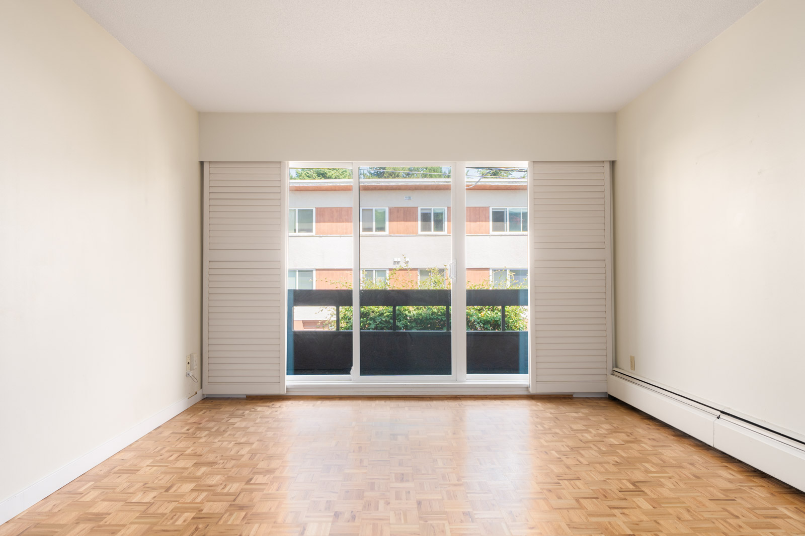 Empty room with light walls, parquet floors, and a large window with sliding shutters facing another building on W 2nd Ave, Vancouver.
