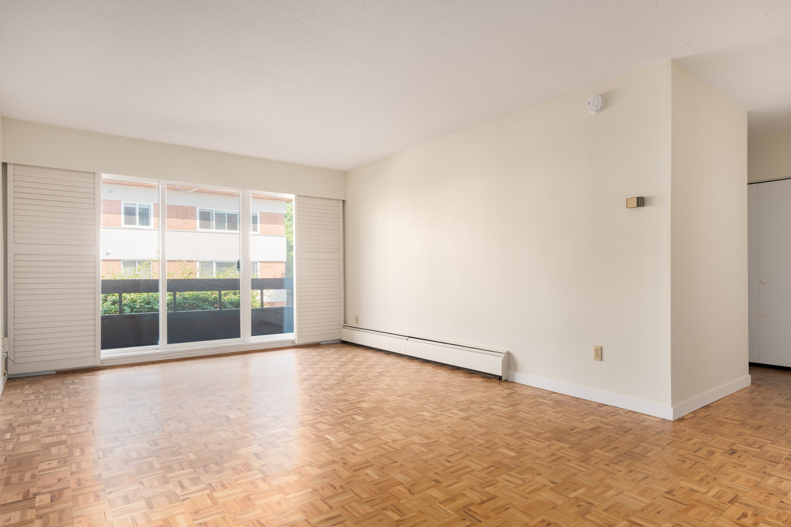 Empty room with parquet floor, large window and shutters, beige walls, baseboard heater, and closet in a Vancouver apartment.