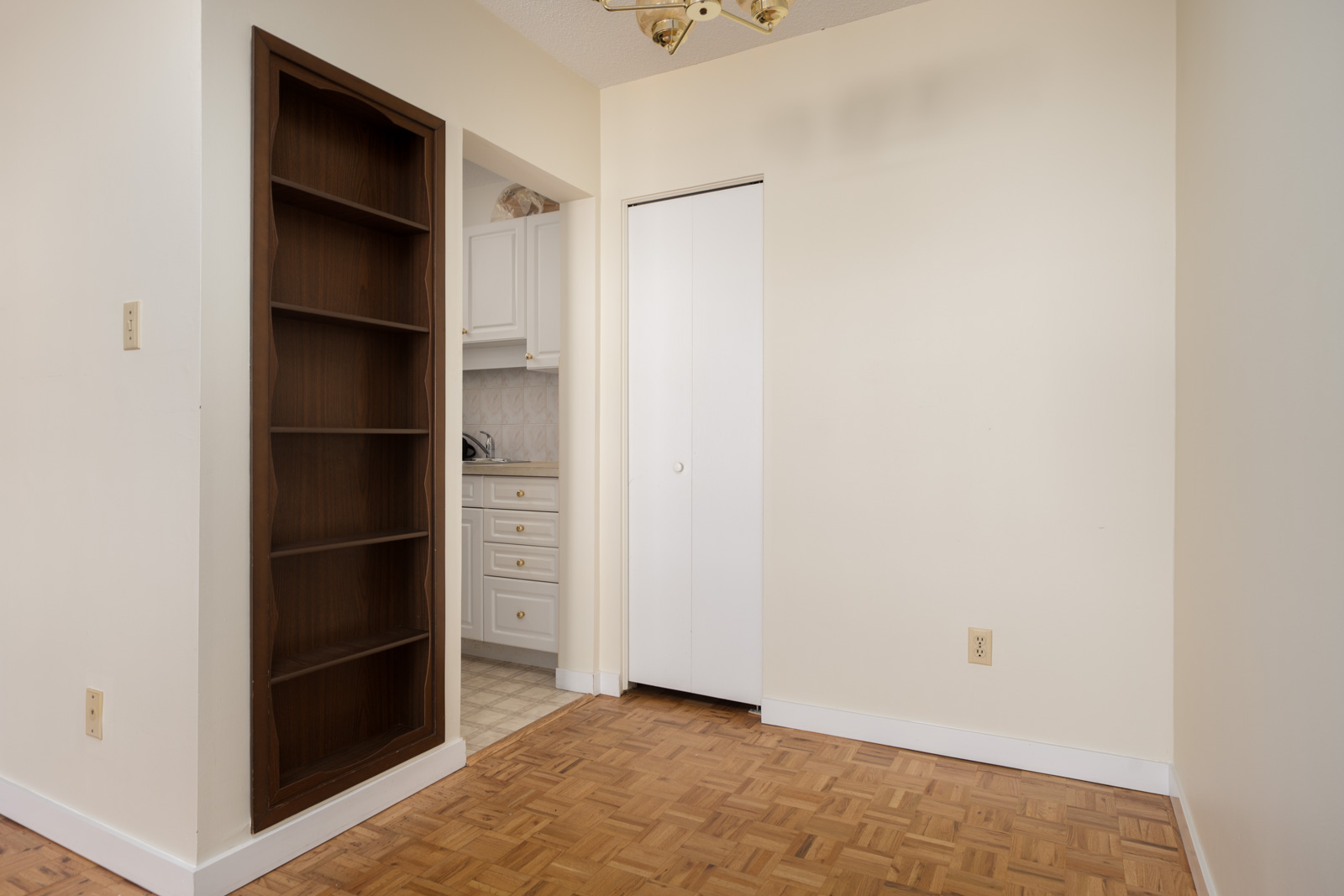 Empty room with parquet floor, built-in brown bookshelf, white closet door, and kitchen view in Vancouver apartment.