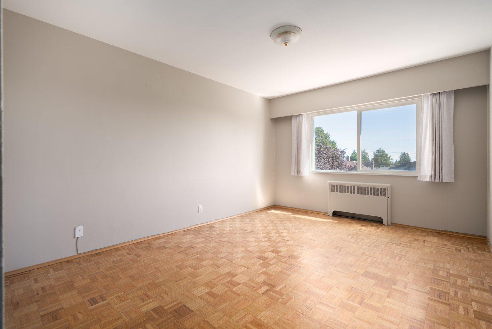 Empty room with light-colored walls, parquet flooring, a ceiling light, and a large window with white curtains letting in daylight.