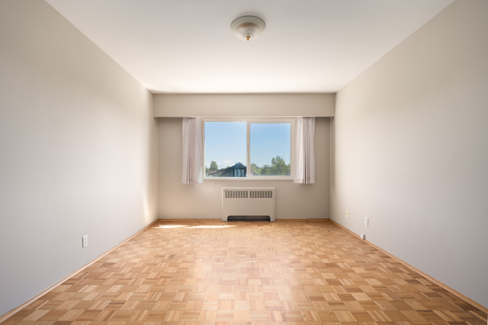 Empty room with light-colored walls, a parquet wood floor, a ceiling light fixture, and a large window with white curtains letting in daylight.