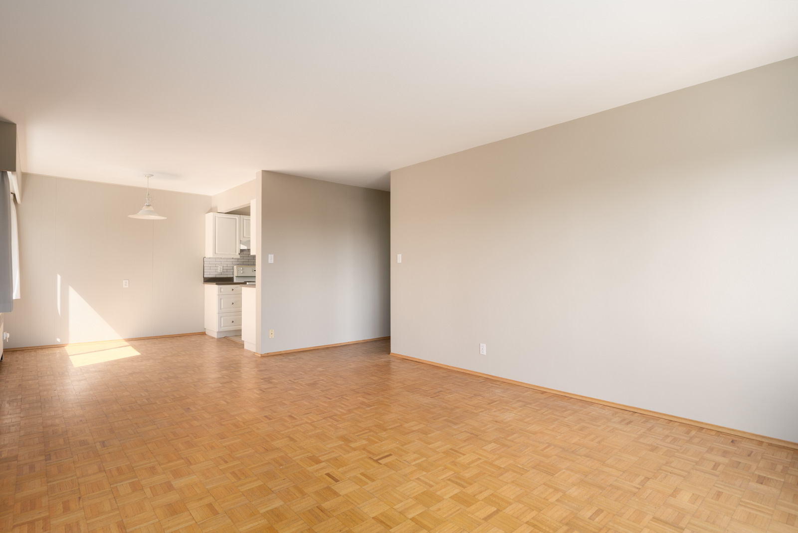 Empty room with light wood parquet flooring, beige walls, and a view of a kitchen area with white cabinets and tile backsplash in the background.