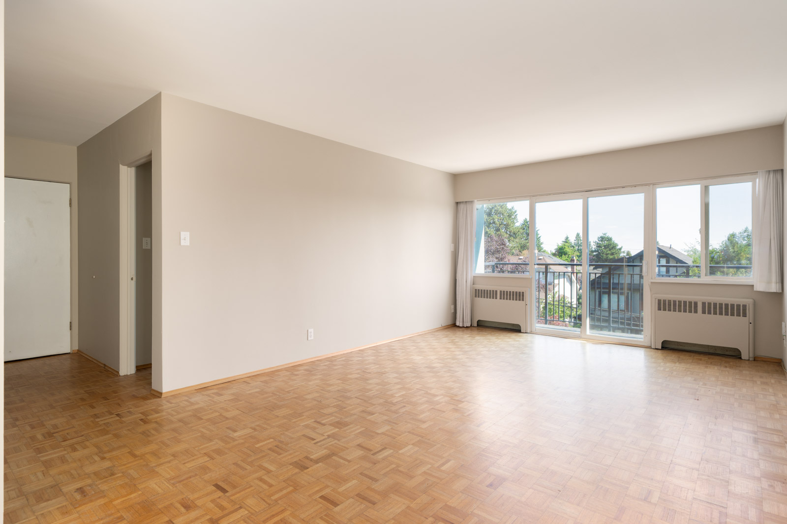 Unfurnished living room with parquet flooring, off-white walls, large window, and glass door leading to a balcony; natural light fills the space.