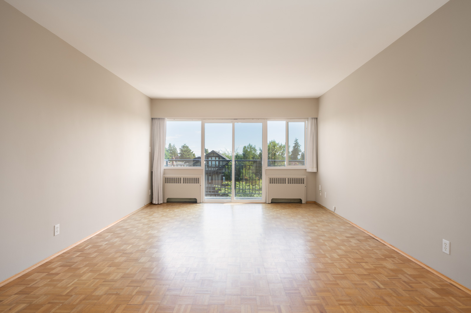 Empty room with light-colored parquet flooring, beige walls, and large windows letting in natural light, overlooking trees and buildings outside.